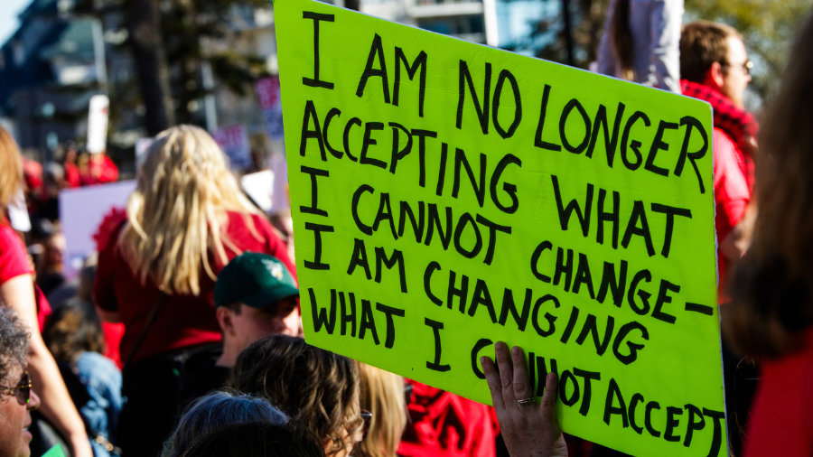 A protesting teacher holding a sign that says I am no longer accepting what I cannot change, I am changing what I cannot accept